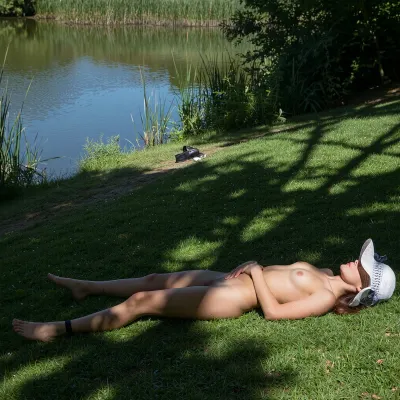 A serene, sun-dappled scene of a woman lying on her back on a grassy bank beside a calm pond. She wears a white sun hat tilted over her face, obscuring her eyes, appearing to rest peacefully. Her arms are crossed over her abdomen, and a black band is visible on her left ankle. The composition places her centrally in the foreground, with dappled sunlight and deep shadows creating a strong contrast across the green grass. In the background, the tranquil water reflects the sky, bordered by tall reeds and lush vegetation. A pair of black shoes and a small white object lie on the grass near the water's edge. The lighting is bright, natural daylight, casting sharp, elongated shadows from unseen trees. The overall atmosphere is one of quiet relaxation in a natural setting. The image has a realistic, photographic style with vibrant colors and a shallow depth of field, focusing sharply on the woman while softly blurring the background.