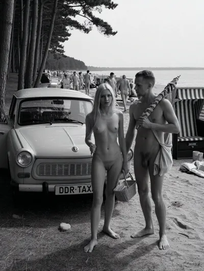 in 1970 on the Baltic Sea coast of the German Democratic Republic, a historically accurate monochrome documentary summer photograph in dramatic East German social realism, high-detail archival black and white photography, a period-correct Trabant parked in the shaded foreground at the edge of a dark coastal pine grove, slightly dusty, plain roof, open passenger door, rectangular front license plate mounted directly on the bumper, clearly reading "DDR-TAXI" in bold uppercase letters, the text integrated like a normal vehicle plate, not a sign, rough sandy ground scattered with pine needles, dry grass, small stones, and tire marks. Beside the car stands an adult young couple, both 25 years old, fully nude in a natural Freikörperkultur context, complete adult nudity shown in a matter-of-fact documentary manner, including his visible penis and her visible breasts, nipples, and pubic area, not erotic and not posed, simply everyday naturist beach life. The woman, long blonde hair, athletic natural figure, calm face, casual posture, locks the Trabant with her right hand using a small key, while a simple wicker beach basket filled with modest food hangs from the crook of her left arm. The man with a relaxed, sporty build carries a folded vintage striped sun umbrella across his shoulders and holds a practical canvas bag with towels. Both move naturally, caught mid-action. Behind them, the pines open toward a busy Baltic naturist beach with many clearly adult vacationers, men and women of different adult ages, all naturally nude, walking, sitting, talking, sunbathing, arranging towels in a casual, respectful atmosphere, several traditional hooded wicker beach chairs with striped fabric and curved canopies, vintage windbreaks, canvas bags, thermos flasks, enamel cups, folded newspapers, soft dunes, pale sand and the calm Baltic Sea. Strong tonal contrast, realistic analog film grain, high texture detail, deep shadows and bright haze, candid composition, respectful historical documentary style, no modern objects, no erotic posing, only authentic 1970s DDR summer life.