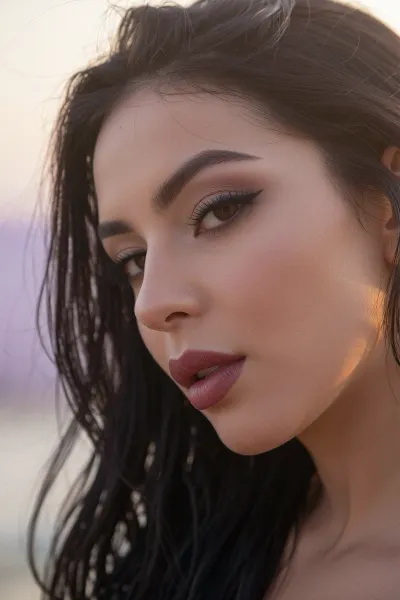 An intimate tight closeup portrait of a sultry Latina woman with an ethereal, soft-focus aesthetic, defined by diffused light. The sensual woman's long elegant dark hair falls loosely in a thick cascade of luxurious hair.
Long feathery eyelashes and dark smokey eye shadow with thick winged eyeliner accentuate her sensual eye gaze and soft brown eyes.
Her thick plump lips are a dark matte berry with pillowy creases. She has high sharp elegant cheekbones and flawless radiant skin.

The scene is enveloped in a powerful but diffused golden-hour glow from her side, which creates a luminous light bloom on her skin, dissolving sharp edges in an ethereal haze. The background is a very blurred, dreamlike pastoral pastel lavender-gray coastal sky, soft and bright. Soft focus on her eyes.
The overall mood is an intimate serene sensuality. Shallow depth of field, subtle film grain, light leaks, vintage analog lens. This is a high-key photograph.
The subject is anchored to the right side of the image, using the rule-of-thirds compositional technique.
The image captures extreme realistic details including sharp limbal rings and fine vellus hairs.