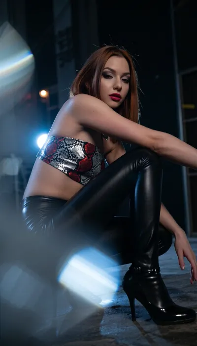 This cinematic portrait captures a attractive petite girl in the style of a distressed photo, The scene depicts a female form in silhouette wearing high heels. The female is squatting on the ground with her elbows resting casually on her knees although the female is only seen in silhouette, black latex leggins, strapless mini top bh with a snakeskin-like pattern in metallic red and silver, hints of color elude to background strobe lights in a gritty urban scene. The focus of the image I’d the contrast between the dark stillness of the silhouette and the intense movement the vivid strobe lights imply, glossy magenta lips and pastel eyeshadow, blurring the foreground into soft out of focus, tactile bokeh, The color grade is melancholic and desaturated, misty atmosphere around her body, glow highlights, bokeh background, prismatic color separation on speculars, strong glow on whole image, girl has dark makeup, redhead hair, bold feline eyeliner, dark red lips, dark grey and silver eyeshadow, golden-amber eyes that create a powerful gaze, fierce hypnotic expression, Deep black background, ultra high quality, cold blue-silver lighting, strong rim light, subtle atmospheric haze, high contrast chiaroscuro, shimmering metallic reflections, mysterious dark fantasy aesthetic, ultra-detailed textures, cinematic photography, shallow depth of field, volumetric lighting, 85mm lens, photorealistic, high fashion editorial, (9:16), vertical composition