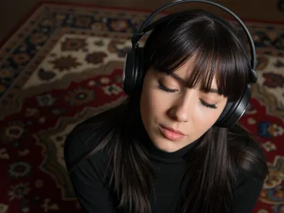 An intimate tight closeup portrait, A young woman, large black headphones on, a Persian rug fills the background, completely absorbed and surrendered to the music, (she has very long dark hair with a fringe, black turtleneck), .


Heavy grain, deep amber lighting, near total darkness, extremely shallow depth of field.
The warm dim light, deep shadow consuming the floor and the edges of everything, the underexposure flattening all contrast into near darkness, the darkness the entire mood.
