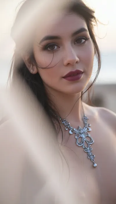A vertical cinematic closeup portrait with an ethereal, soft-focus aesthetic, defined by diffused light. The subject is an enigmatic young Latina woman with long dark hair and wispy strands that have a soft luminous halo framing her elegant facial features with stray strands of windblown hair. She wears dark smoky makeup that highlights her large expressive eyes, while her soft full lips are a deep matte berry. Her elegant neckline is accented by an intricate antique silver gothic necklace. The soft lighting and shadows flatter her slender body. The scene is enveloped in a powerful but diffused golden-hour glow from the side, which creates a luminous light bloom on her skin and the fabric, dissolving sharp edges in an ethereal haze. The background is a very blurred, dreamlike pastoral pastel lavender-gray coastal sky, soft and bright. The overall mood is serene contentment. Shallow depth of field, subtle film grain. This is a high-key photograph.
