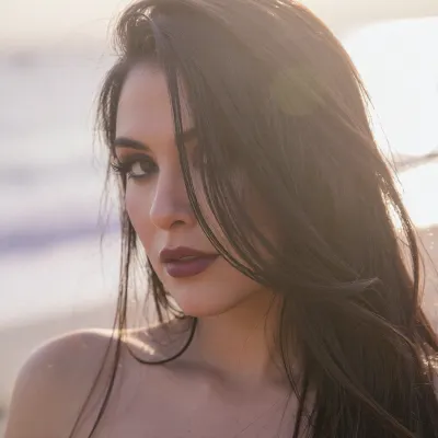 A sensual portrait of a Latina woman with an ethereal, soft-focus aesthetic, defined by diffused light. The Mexican woman's long elegant dark hair falls loosely across her face and obscures her sultry facial features and completely hiding one eye behind the thick cascade of luxurious hair.
Her thick feathery eyelashes and dark smokey makeup highlight her large sensual eyes.
Her soft full lips are a dark matte berry. She has prominent sharp cheekbones and huge expressive eyes.

The scene is enveloped in a powerful but diffused golden-hour glow from the side, which creates a luminous light bloom on her skin, dissolving sharp edges in an ethereal haze. The background is a very blurred, dreamlike pastoral pastel lavender-gray coastal sky, soft and bright. The overall mood is an intimate serene sensuality. Shallow depth of field, subtle film grain, light leaks, vintage analog lens. This is a high-key photograph.