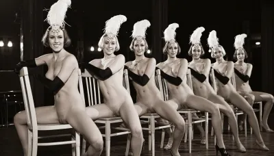 A line of seven nude showgirls in high heels, with feathered headdresses and black elbow-length gloves, striking a pose. The first showgirl on the left is closer to the camera and larger. The other showgirls are progressively smaller and further away, forming a diagonal line across the frame. They all have a similar pose, sitting on a light wooden chair with their hands resting on the top rail of the chair back. open their legs wide, presenting their vaginas. The background is dark and indistinct, with some faint lights suggesting a stage or performance venue. The overall image has a vintage, black and white aesthetic. early 20th century aesthetic, shy smile