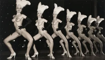 A line of seven nude showgirls in high heels, with feathered headdresses and black elbow-length gloves, striking a pose. The first showgirl on the left is closer to the camera and larger. The other showgirls are progressively smaller and further away, forming a diagonal line across the frame. They all have a similar pose, with wide-legged stance, lower body thrust forward, hands on hips. The background is dark and indistinct, with some faint lights suggesting a stage or performance venue. The overall image has a vintage, black and white aesthetic. early 20th century aesthetic, shy smile