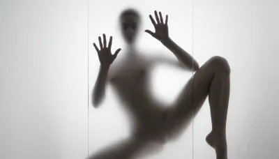 Ultra photorealistic minimalist monochrome fine art photography, vertical composition, mysterious adult female silhouette 28 years old standing behind heavily frosted translucent glass, entire image dominated by luminous white and pale gray tones, figure positioned almost exactly in the center, body visible only as a soft charcoal shadow through condensation and diffused glass texture, no facial detail readable, head slightly inclined, shoulders narrow, torso softly defined, natural feminine proportions, blurred hip and waist transitions, absolutely no sharp body edges, both arms raised forward toward the glass, both flat inner palms pressed firmly against the frosted pane, fingers naturally spread, the two hand contact areas are the only elements in the entire image rendered razor sharp, crisply outlined, perfectly defined and hyperdetailed, individual fingers, fingertips, palm creases, subtle skin texture, fine lines, pressure points and delicate condensation displacement around each hand clearly visible, faint moisture halos surrounding the palms, realistic contact between warm skin and cold glass, while wrists, arms, body, head and legs immediately dissolve back into heavy diffusion and soft blur, thick atmospheric frosting obscuring all other anatomical detail, one leg straight and weight bearing, the other knee bent inward creating an elegant asymmetrical S shaped silhouette, brightest values surrounding the figure and gradually fading toward the center, extremely soft optical blooming, subtle vertical streaks and uneven translucency across the glass, barely perceptible shadows around the feet, dreamy ethereal isolation, no visible room details, no hard light source, broad diffuse backlighting from behind the glass, very low contrast, milky whites, pearl gray midtones and soft graphite silhouette, strong selective focus exclusively on the palms against the pane, everything else intentionally defocused, slight vintage film softness, minimal grain, 85 mm lens compression, shallow tonal depth, haunting anonymous presence, museum quality abstract photographic realism