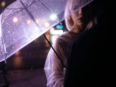 Close-up on her face, she faces the viewer, A highlight on her face, her face is visible in the darkness, A partial silhouette of a woman standing, blond hair in a bob cut, breasts, she wears a clear plastic rain coat, holding an open umbrella, rain rendered as glowing violet-purple light trails streaking downward off the umbrella edge, soft wet-glass texture with sharp raindrop streaks in the near foreground, dreamy out-of-focus bokeh lights in warm pink, orange and cool teal glowing softly behind, wet reflective ground, long exposure light painting style, high contrast black and purple palette with soft romantic bokeh glow, moody night atmosphere.