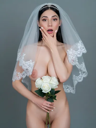 A full-body studio portrait of a gorgeous model, (in her late 20s, with long dark hair, a perfect figure, and large breasts), standing nude against a plain, cool grey background. She is wearing a sheer white lace-edged bridal veil and a delicate silver tiara, holding a small bouquet of white roses in one hand while her other hand rests on her cheek in a surprised gesture, o. She holds the white rose bouquet against her pubic area, obscuring her genitalia while leaving the rest of her naked female body completely bare. The lighting is soft and diffused, highlighting her pale skin and the transparency of the tulle veil. The composition includes a close-up crop focusing on her face, the texture of the veil, and her breast, emphasizing the contrast between the innocent bridal accessories and her stark nudity. Her nails are manicured with a pale white polish, and the overall aesthetic is sensual, erotic, and reminiscent of high-key glamour photography.