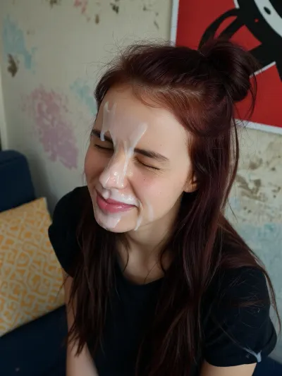 A young tall very skinny woman with long, dark reddish-brown hair, styled in a messy bun on top of her head with loose strands falling around her shoulders, her face covered in a white, viscous liquid. Her eyes are tightly squeezed shut, and her mouth is slightly open, revealing a hint of pink lips also covered in the liquid. She has a flushed complexion with some visible skin texture and small blemishes. She is wearing a dark, possibly black, t-shirt.

The background features a textured wall with subtle, faded pastel colors like light blue, pink, and beige, suggesting a worn or artistic finish. In the upper right corner, a portion of a vibrant red poster or artwork with abstract black patterns is visible. To the left, a yellow patterned pillow rests on a dark blue surface, possibly a bed or couch.

The lighting is direct and somewhat harsh, casting minimal shadows and highlighting the texture of the liquid on her face. The overall mood is intense and raw.

Captured with a close-up, slightly high-angle shot, using a wide aperture for a shallow depth of field, focusing sharply on her face. The image has a slightly grainy, candid, and unrefined aesthetic, reminiscent of early digital photography, with natural color grading.