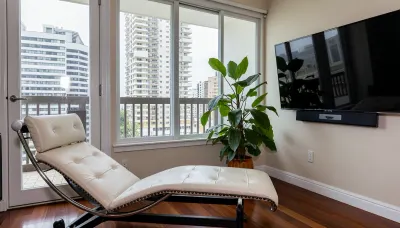 The scene is a high-rise apartment, skyscraper, large floor to ceiling windows, aged mahogany floors, a cream colored leather chaise lounge, a large Epipremnum aureum, money plant, a 75 inch TV is on the wall, a balcony.