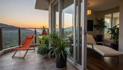 The scene, looking out at the sunset on the corner balcony of a 12 ft ceiling apartment, large floor to ceiling windows, skyscraper, stone floors on the outside, mahogany wood floors on the inside, a cream colored lounge chair on the inside, a burnt-ornange deck chair on the balcony. SEVERAL PLANTS are on the inside and outside of apartment, [types of plants include a matured mix of the following], (Epipremnum aureum), (Dracaena trifasciata), (Philodendron scandens), (Hedera helix), far in the distance is a mountain that is deeply forested........there is a helipad