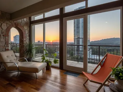 The scene, looking out at the sunset on the corner balcony of a 12 ft ceiling apartment, large floor to ceiling windows, skyscraper, focus on the outside, while balcony has stone floors, the interior which is an open floorplan with aged mahogany wood floors, a cream colored lounge chair is on the inside, a burnt-ornange deck chair can be found on the outside, large ceiling beam goes across the ceiling, stone arch, brick walls, SEVERAL MATURED PLANTS are on the inside and outside of the apartment, [types of plants include a matured mix of the following], (Epipremnum aureum), (Dracaena trifasciata), (Philodendron scandens), (Hedera helix), far in the distance is a mountain that is deeply forested, one of the plants hangs from the ceiling. The balcony is half ceiling half skylight.