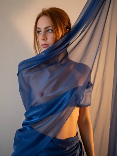 A woman standing against a wall, wrapped in a translucent blue-blue, flowing cloth. The woman has redhead hair and looks thoughtful, deep green eyes, and the fabric reveals a part of her body, creating spectacular and intriguing lighting. The decor seems minimalistic, with a warm, golden light that highlights the drapery of the fabric and the figure of the model. The focus is on the interaction of the figure and the fluttering fabric.