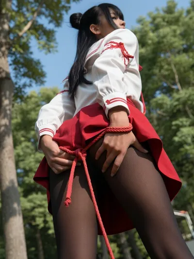 Low-angle shot from behind, a woman wearing a white long-sleeved blouse with red trim and traditional red lacing, a short layered red skirt, and opaque black tights, red cords tightly binding her wrists together behind her back, with several visible knots and loops. She bends forward slightly, holding the back of her skirt with both hands, legs close together and raised behind her. Long black hair tied up in a bun. Outdoor setting with lush green trees and blue sky, bright natural sunlight, soft background blur, cinematic perspective, realistic fabric folds, detailed clothing, shallow depth of field, vertical composition, photorealistic.