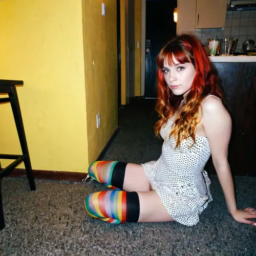 (A 22-year-old introvert redhead grunge girl with bangs and multi-coloured stockings and a cute short cotton dress is sitting on the floor in a dark corner drinking lemonade from a glass at a party in 1997), (an older 27-year-old tall and slim woman with long wavy dark blonde hair is barely visible in the background), photo-realistic, compact camera shot, analogue film artifacts, grainy, long shot, [[naked]]