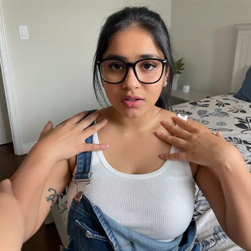 From first person POV perspective. You are alone in your bedroom with a classmate named Priya. She's a cute chubby south indian girl with a busty curvy body. Her Dark brown melanin rich skin stands in contrast to your white skin and fair complexion. She's slightly nerdy and into alternative music. You stand near her as she sits on your bed. Your hands are reaching down to caress her shoulder and upper body. She looks a little shocked and worried. Priya thought you invited her over to study. She wears overalls and a white ribbed tank top. Black frame glasses and her hair tied back. She has a few simple tattoos.