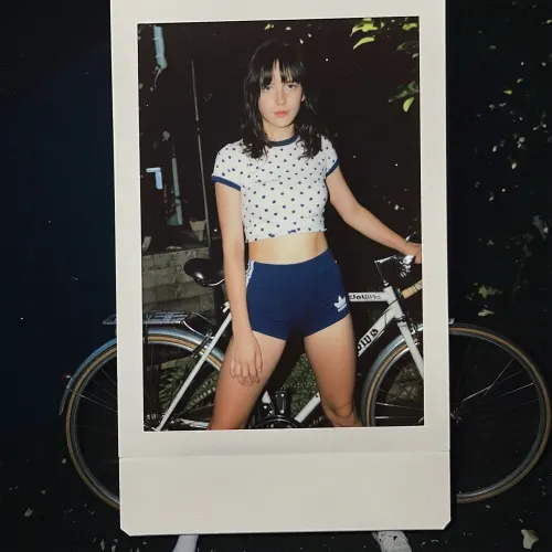 Polaroid, bent damaged Polaroid, sun faded image, bad quality photo, camera flash, slightly dark background. 1980s grainy bad quality colour photo, slightly dark background. 29-year-old italian girl messy black bangs shoulder length hair.thin figure, extremely wide hips, White polka dot t-shirt, navy blue 1980s adidas terry cloth volley-ball shorts, short white socks, sneekers. oval face, small nose, high cheek bones, narrow mouth. backyard summer, standing beside her sekine 10-speed bicycle.
