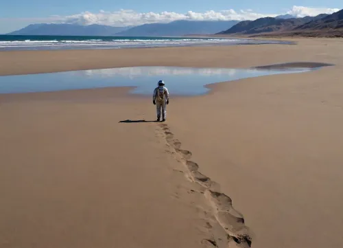 each color is solid and separated from the other by a thin line of a darker shade, bright yellow sand beach with the dunes which contours outlined by dark brown lines in the foreground flows into the soft shapes of the high mountains in the background, their contours outlined by dark grey lines, sky is dark blue, turning into purple in the distance, with red reflections over the mountains, it must be evening, aquamarine of the reservoir complete width of the field of view separates the beach from the mountains on the horizon, lonely small figure of a human in bright silver metal spacesuit with black face shield stands in foreground on the beach, a trail of footprints stretches across the sand from the human to the edge of the image, laconism and graphicity in the drawing of landscape evoke poster in constructivism style and enhance the dramatic effect, is enhanced by the light, which gives almost no shadows, the image is a symbol of loneliness and lostness in space