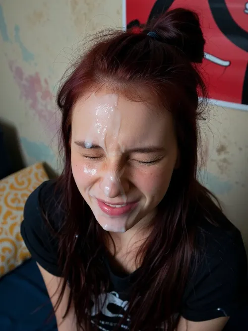 A young woman with long, dark reddish-brown hair, styled in a messy bun on top of her head with loose strands falling around her shoulders, her face covered in a white, viscous liquid. Her eyes are tightly squeezed shut, and her mouth is slightly open, revealing a hint of pink lips also covered in the liquid. She has a flushed complexion with some visible skin texture and small blemishes. She is wearing a dark, possibly black, t-shirt.

The background features a textured wall with subtle, faded pastel colors like light blue, pink, and beige, suggesting a worn or artistic finish. In the upper right corner, a portion of a vibrant red poster or artwork with abstract black patterns is visible. To the left, a yellow patterned pillow rests on a dark blue surface, possibly a bed or couch.

The lighting is direct and somewhat harsh, casting minimal shadows and highlighting the texture of the liquid on her face. The overall mood is intense and raw.

Captured with a close-up, slightly high-angle shot, using a wide aperture for a shallow depth of field, focusing sharply on her face. The image has a slightly grainy, candid, and unrefined aesthetic, reminiscent of early digital photography, with natural color grading.