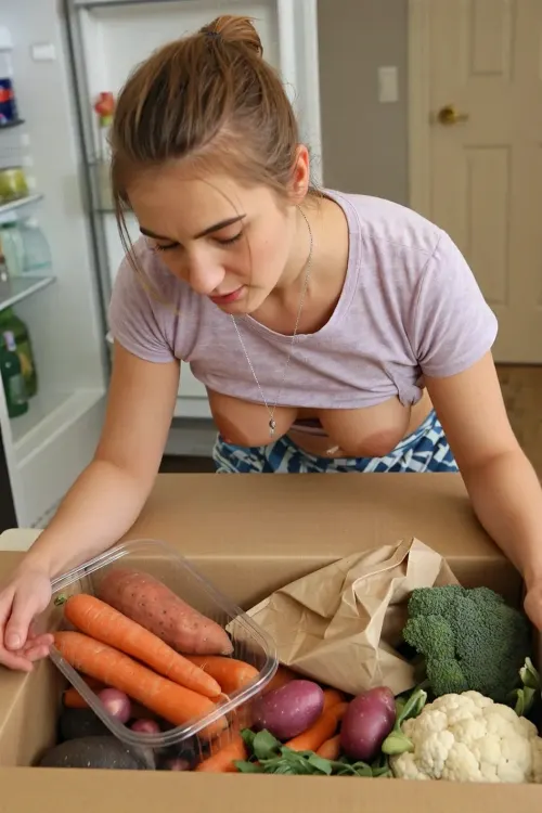 A woman with light brown hair pulled back in a messy bun, wearing a light purple patterned t-shirt and a silver chain necklace with a small pendant, is bending over, revealing a glimpse of a patterned blue garment underneath and her tits. She is looking down into a box. Her right arm is visible, holding a clear plastic container filled with vegetables including orange carrots, reddish-orange sweet potatoes, and small purple potatoes, which she is placing into a larger cardboard box. Inside the cardboard box, other vegetables like carrots, purple potatoes, broccoli, and cauliflower are visible, along with a crumpled brown paper bag.

The scene is set indoors, likely in a kitchen, with a refrigerator partially visible in the background on the left, containing bottles and containers. A light-colored door is visible in the upper right background.

The lighting is bright and even, illuminating the woman and the vegetables clearly. The colors are natural and vibrant, emphasizing the freshness of the produce.

The shot is a medium close-up, taken from a slightly elevated angle, looking down at the woman and the boxes. The composition focuses on the action of placing the vegetables, with a shallow depth of field that keeps the woman and the immediate foreground in sharp focus.

The overall style is realistic and candid, resembling a snapshot or documentary photograph. High resolution, sharp focus, natural color grading.