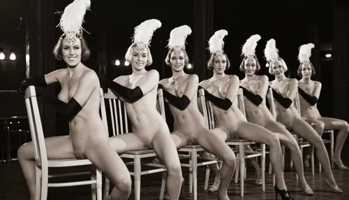 A line of seven nude showgirls in high heels, with feathered headdresses and black elbow-length gloves, striking a pose. The first showgirl on the left is closer to the camera and larger. The other showgirls are progressively smaller and further away, forming a diagonal line across the frame. They all have a similar pose, sitting on a light wooden chair with their hands resting on the top rail of the chair back. open their legs wide, presenting their vaginas. The background is dark and indistinct, with some faint lights suggesting a stage or performance venue. The overall image has a vintage, black and white aesthetic. early 20th century aesthetic, shy smile