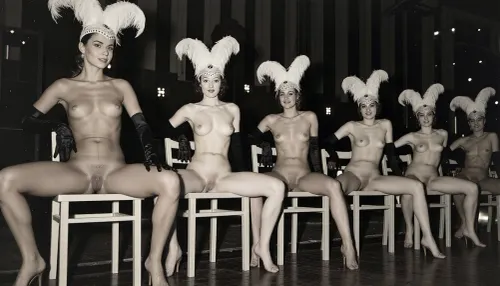 A line of seven nude showgirls in high heels, with feathered headdresses and black elbow-length gloves, striking a pose. The first showgirl on the left is closer to the camera and larger. The other showgirls are progressively smaller and further away, forming a diagonal line across the frame. They all have a similar pose, sitting on a light wooden chair with their hands resting on the top rail of the chair back. open their legs wide, presenting their vaginas. The background is dark and indistinct, with some faint lights suggesting a stage or performance venue. The overall image has a vintage, black and white aesthetic. early 20th century aesthetic, shy smile