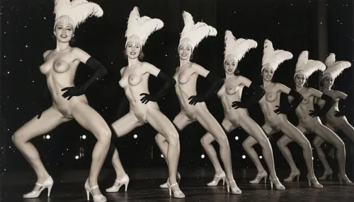 A line of seven nude showgirls in high heels, with feathered headdresses and black elbow-length gloves, striking a pose. The first showgirl on the left is closer to the camera and larger. The other showgirls are progressively smaller and further away, forming a diagonal line across the frame. They all have a similar pose, with wide-legged stance, lower body thrust forward, hands on hips. The background is dark and indistinct, with some faint lights suggesting a stage or performance venue. The overall image has a vintage, black and white aesthetic. early 20th century aesthetic, shy smile