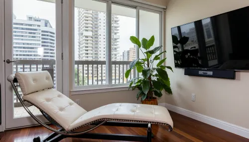The scene is a high-rise apartment, skyscraper, large floor to ceiling windows, aged mahogany floors, a cream colored leather chaise lounge, a large Epipremnum aureum, money plant, a 75 inch TV is on the wall, a balcony.