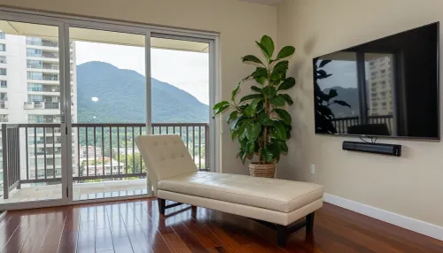 The scene is a high-rise apartment, skyscraper, large floor to ceiling windows, aged mahogany floors, a cream colored leather chaise lounge, a large Epipremnum aureum, money plant, a 75 inch TV is on the wall, a balcony, far in the distance is a mountain that is deeply forested.