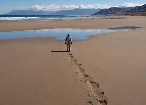 each color is solid and separated from the other by a thin line of a darker shade, bright yellow sand beach with the dunes which contours outlined by dark brown lines in the foreground flows into the soft shapes of the high mountains in the background, their contours outlined by dark grey lines, sky is dark blue, turning into purple in the distance, with red reflections over the mountains, it must be evening, aquamarine of the reservoir complete width of the field of view separates the beach from the mountains on the horizon, lonely small figure of a human in bright silver metal spacesuit with black face shield stands in foreground on the beach, a trail of footprints stretches across the sand from the human to the edge of the image, laconism and graphicity in the drawing of landscape evoke poster in constructivism style and enhance the dramatic effect, is enhanced by the light, which gives almost no shadows, the image is a symbol of loneliness and lostness in space