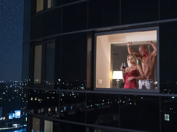 A striking nocturnal photograph captures the facade of a modern luxury hotel, its dark glass exterior reflecting the starless night sky and distant city lights. The camera focuses intently on a single, large rectangular window situated on the third floor, which serves as the sole point of illumination against the surrounding darkness. Inside this window, a woman stands silhouetted by the warm glow of an interior lamp, her figure framed perfectly by the glass pane. She is a stunning blonde woman in her early thirties standing exquisitely poised. She is clad in rich, vibrant red lingerie that complements her sophisticated presence, creating a striking contrast against the cool, dark tones of the night. Her face is made up exquisitely, red lips & eyeshadow to compliment her blue eyes. She cradles a stemmed glass of deep red wine. Her skin glows with an inner warmth, illuminated by the bright light spilling from within the room behind her, casting soft shadows across the glass and highlighting the intricate details of her attire, creating a soft, glowing outline against the deep shadows of the room. Her posture suggests a moment of quiet contemplation or longing as she gazes out into the cool, silent night, her face partially obscured but her presence commanding the viewer's attention through the barrier of the window. Behind her is a handsome, muscular man, naked except for striped shorts. He is bound and gagged with a red gag and chained to the wall, his hands fastened above his head with cuffs. The lighting is dramatic and cinematic, utilising high-contrast chiaroscuro to emphasize the separation between the intimate warmth of the interior and the cold isolation of the exterior. The texture of the glass is rendered with realistic reflections and refractions, adding depth and complexity to the composition while maintaining a sense of distance and mystery.

The image employs a shallow depth of field to keep the woman and man sharply in focus while blurring the hotel's architectural details in the background, enhancing the emotional intimacy of the scene. The composition is centered and symmetrical, reinforcing the feeling of stillness and solitude. The mood is atmospheric and evocative, blending elements of romance and melancholy, inviting the viewer to wonder about the story behind the woman's gaze. The overall aesthetic is polished and professional, capturing the essence of urban loneliness and personal reflection through the medium of high-fidelity photography.