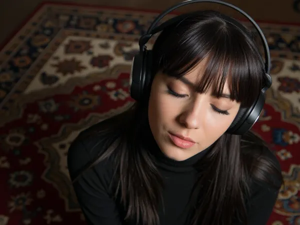 An intimate tight closeup portrait, A young woman, large black headphones on, a Persian rug fills the background, completely absorbed and surrendered to the music, (she has very long dark hair with a fringe, black turtleneck), .


Heavy grain, deep amber lighting, near total darkness, extremely shallow depth of field.
The warm dim light, deep shadow consuming the floor and the edges of everything, the underexposure flattening all contrast into near darkness, the darkness the entire mood.