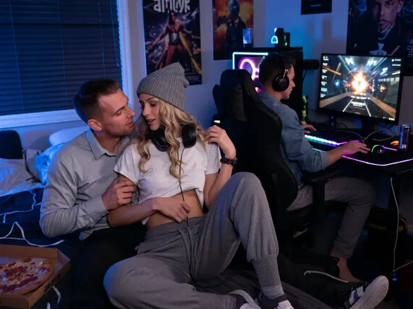 Ultra low-key cinematic gaming room portrait, computer monitor as dominant key light, deep near-black shadows, rich OLED-like contrast, subtle blue and purple RGB rim light, deliberately underexposed by about two stops, 35 mm full-frame documentary viewpoint, f/1.4 shallow depth of field, ISO 800, subtle analog film grain, strong micro-contrast, photorealistic realism, dark messy late-night gamer bedroom with lowered blinds, colorful LED strips, glowing RGB gaming setup, illuminated mechanical keyboard, mouse, controller, headset cables, pizza box, energy drink cans, tangled cables and fictional open-world action game posters, exactly three adults arranged in two clearly separated depth planes, a couple has sex in the foreground on the bed, an adult gamer woman, 25 years old, and her husband, 38 years old, they are married, affectionate and visibly devoted to each other, being close and intimate together on the rumpled bed, warm eye contact, subtle touching, relaxed playful smiles and intimate familiar chemistry, the woman has long blonde corkscrew curls beneath a small knitted wool beanie, gaming headset around her neck, toned athletic figure, warm tan skin, cropped loose T-shirt revealing her midriff, thick gray sweatpants and warm wool socks, her husband looks comparatively conservative and understated, neatly groomed slightly gray hair, clean casual shirt, dark trousers, calm friendly appearance, behind the couple, clearly separated in the background, another adult man, 28 years old, sits alone in a black gaming chair at the gaming desk, wearing a headset, leaning toward the large monitor, eyes fixed on the game, both hands on the illuminated keyboard and mouse, fully focused on gaming, the monitor displays a fictional neon open-world action game, monitor glow illuminating the room, foreground married couple dominant and sharp, background gamer visibly separate, natural depth of field, immersive cinematic photorealism