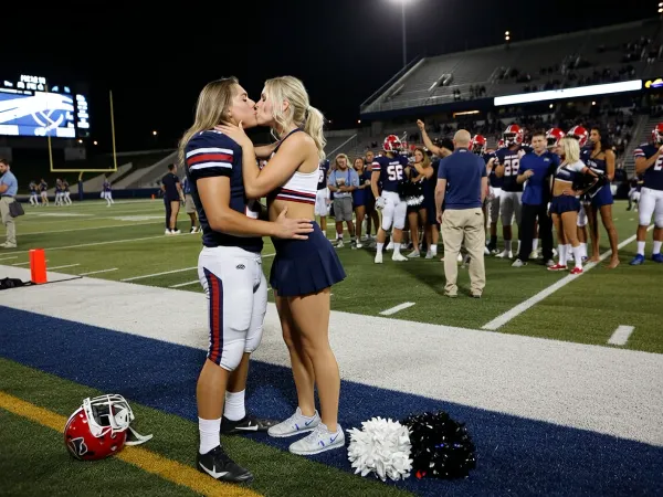 A cinematic, photorealistic late-night scene immediately after an American football game, depicting two adult women sharing a spontaneous romantic kiss at the edge of the football field after the final whistle.

The main subjects are unmistakably TWO WOMEN, a curvy feminine beautiful female American football quarterback and a voluptuous female cheerleader, both in their mid-twenties. The female quarterback has just led her team to victory after an intense game. She is clearly and visibly a young woman, with an athletic feminine build, feminine facial features, and long slightly messy blonde hair. She is still wearing her complete American football uniform with padded pants, shoulder pads, team jersey, and cleats, but her helmet has been removed and is discarded on the ground.

Overcome with happiness after the win, the female quarterback has finally acted on the feelings she has secretly had for the cheerleader throughout the entire season. She stands face-to-face with the cheerleader and they are kissing passionately but tenderly, the cheerleader holding one hand against the quarterback's cheek while the quarterback has an arm around her waist. Their body language should feel spontaneous, emotional, affectionate, and slightly overwhelmed, like a long-awaited moment finally happening.

The cheerleader is also unmistakably an adult woman in her mid-twenties, attractive and athletic, wearing a realistic professional American football cheerleading uniform consisting of a cropped top and pleated skirt, with team colors, athletic shoes, and her platinum blond hair styled in a ponytail for the game. A pair of pom-poms lies forgotten on the turf beside them.

They stand near the sideline at the edge of the football field. Behind them, slightly out of focus but clearly recognizable, the quarterback's teammates are celebrating the victory, some still wearing helmets, others embracing, shouting, or raising their arms. Coaches, staff, cheerleaders, photographers, and spectators remain visible in the background. The stadium stands are filled with fans beginning to leave while others continue celebrating.

Bright stadium floodlights illuminate the field against the dark night sky. The turf is worn from the game, with painted yard lines, sideline markings, scattered equipment, and distant goalposts visible. The scoreboard in the background suggests that the home team has narrowly won.

Focus strongly on the two adult women and their emotional kiss while retaining enough environmental detail to clearly establish the aftermath of a major American football game. Shallow depth of field, cinematic stadium lighting, realistic skin texture, realistic athletic proportions, natural facial expressions, believable football equipment, subtle sweat and grass stains on the quarterback's uniform, dramatic sports photography aesthetic, emotionally intimate moment within a large celebratory stadium environment, high detail, natural anatomy, photorealistic.