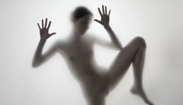 Ultra photorealistic minimalist monochrome fine art photography, vertical composition, mysterious adult female silhouette 28 years old standing behind heavily frosted translucent glass, entire image dominated by luminous white and pale gray tones, figure positioned almost exactly in the center, body visible only as a soft charcoal shadow through condensation and diffused glass texture, no facial detail readable, head slightly inclined, shoulders narrow, torso softly defined, natural feminine proportions, blurred hip and waist transitions, absolutely no sharp body edges, both arms raised forward toward the glass, both flat inner palms pressed firmly against the frosted pane, fingers naturally spread, the two hand contact areas are the only elements in the entire image rendered razor sharp, crisply outlined, perfectly defined and hyperdetailed, individual fingers, fingertips, palm creases, subtle skin texture, fine lines, pressure points and delicate condensation displacement around each hand clearly visible, faint moisture halos surrounding the palms, realistic contact between warm skin and cold glass, while wrists, arms, body, head and legs immediately dissolve back into heavy diffusion and soft blur, thick atmospheric frosting obscuring all other anatomical detail, one leg straight and weight bearing, the other knee bent inward creating an elegant asymmetrical S shaped silhouette, brightest values surrounding the figure and gradually fading toward the center, extremely soft optical blooming, subtle vertical streaks and uneven translucency across the glass, barely perceptible shadows around the feet, dreamy ethereal isolation, no visible room details, no hard light source, broad diffuse backlighting from behind the glass, very low contrast, milky whites, pearl gray midtones and soft graphite silhouette, strong selective focus exclusively on the palms against the pane, everything else intentionally defocused, slight vintage film softness, minimal grain, 85 mm lens compression, shallow tonal depth, haunting anonymous presence, museum quality abstract photographic realism