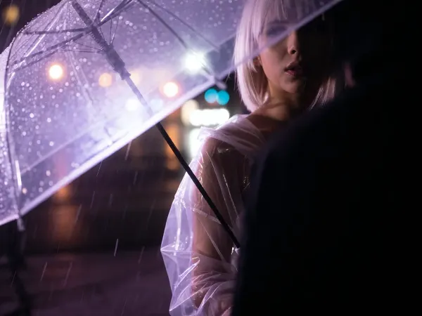 Close-up on her face, she faces the viewer, A highlight on her face, her face is visible in the darkness, A partial silhouette of a woman standing, blond hair in a bob cut, breasts, she wears a clear plastic rain coat, holding an open umbrella, rain rendered as glowing violet-purple light trails streaking downward off the umbrella edge, soft wet-glass texture with sharp raindrop streaks in the near foreground, dreamy out-of-focus bokeh lights in warm pink, orange and cool teal glowing softly behind, wet reflective ground, long exposure light painting style, high contrast black and purple palette with soft romantic bokeh glow, moody night atmosphere.