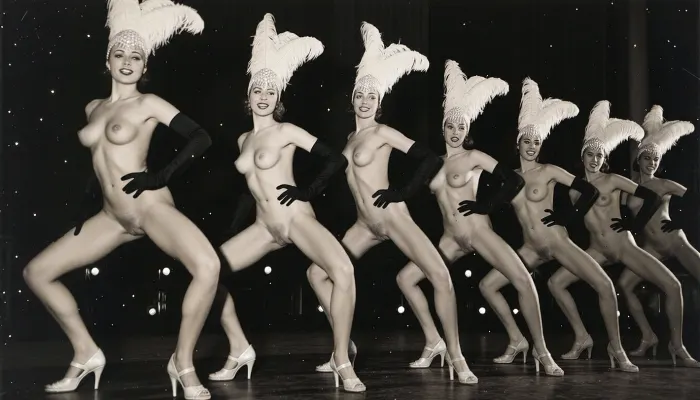 A line of seven nude showgirls in high heels, with feathered headdresses and black elbow-length gloves, striking a pose. The first showgirl on the left is closer to the camera and larger. The other showgirls are progressively smaller and further away, forming a diagonal line across the frame. They all have a similar pose, with wide-legged stance, lower body thrust forward, hands on hips. The background is dark and indistinct, with some faint lights suggesting a stage or performance venue. The overall image has a vintage, black and white aesthetic. early 20th century aesthetic, shy smile