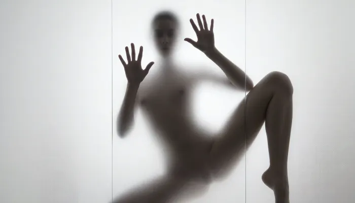Ultra photorealistic minimalist monochrome fine art photography, vertical composition, mysterious adult female silhouette 28 years old standing behind heavily frosted translucent glass, entire image dominated by luminous white and pale gray tones, figure positioned almost exactly in the center, body visible only as a soft charcoal shadow through condensation and diffused glass texture, no facial detail readable, head slightly inclined, shoulders narrow, torso softly defined, natural feminine proportions, blurred hip and waist transitions, absolutely no sharp body edges, both arms raised forward toward the glass, both flat inner palms pressed firmly against the frosted pane, fingers naturally spread, the two hand contact areas are the only elements in the entire image rendered razor sharp, crisply outlined, perfectly defined and hyperdetailed, individual fingers, fingertips, palm creases, subtle skin texture, fine lines, pressure points and delicate condensation displacement around each hand clearly visible, faint moisture halos surrounding the palms, realistic contact between warm skin and cold glass, while wrists, arms, body, head and legs immediately dissolve back into heavy diffusion and soft blur, thick atmospheric frosting obscuring all other anatomical detail, one leg straight and weight bearing, the other knee bent inward creating an elegant asymmetrical S shaped silhouette, brightest values surrounding the figure and gradually fading toward the center, extremely soft optical blooming, subtle vertical streaks and uneven translucency across the glass, barely perceptible shadows around the feet, dreamy ethereal isolation, no visible room details, no hard light source, broad diffuse backlighting from behind the glass, very low contrast, milky whites, pearl gray midtones and soft graphite silhouette, strong selective focus exclusively on the palms against the pane, everything else intentionally defocused, slight vintage film softness, minimal grain, 85 mm lens compression, shallow tonal depth, haunting anonymous presence, museum quality abstract photographic realism