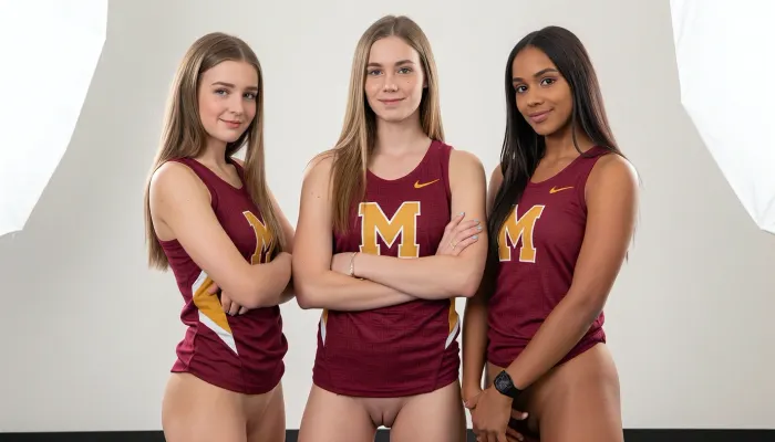A realistic studio photograph of three nude young female athletes standing side by side against a seamless white backdrop, posed only in matching maroon and gold collegiate track and field jersey. all three athletes with perfect shaved vaginas, completely hairless vaginas all visible. The image is captured straight-on with a medium telephoto lens around 85mm at f/8, keeping all three subjects in sharp focus with minimal depth of field falloff, lit by a soft even two-light studio setup with large diffused softboxes positioned front-left and front-right, producing flat even illumination with very soft shadows beneath the chin and arms. The leftmost woman stands angled slightly toward the center, her arms folded across her chest, her body turned in three-quarter view, with long straight blonde hair falling past her shoulders, fair skin with light freckling, sweet shy smile, light brown eyebrows, and pale blue eyes looking directly at the camera, wearing a thin gold bracelet on her left wrist. The center woman stands squarely facing the lens, arms crossed over her chest, with long straight honey-blonde hair parted in the middle framing her face, fair skin, light blue-grey eyes, a sweet shy smile, wearing a silver bracelet and a gold ring on her right hand with pale blue nail polish visible on her fingers.

The rightmost woman stands with her body turned slightly toward the center but her face angled to camera, arms folded with her left hand resting on her right forearm, revealing a black sports watch with silver buckle on her left wrist, long straight dark blonde hair framing her face, warm olive-toned fair skin, brown eyes, and a sweet shy smile. All three only wear identical maroon sleeveless athletic tank tops featuring an allover faint geometric Greek key pattern woven into the fabric, with a large gold interlocking M logo centered on the chest, a small gold Nike swoosh on the upper right chest. The fabric shows a matte synthetic sheen typical of competition uniforms, with visible seam lines and a slight stretch across the torso. The background is pure seamless white with a faint grey gradient at the floor line, no props or environmental elements, the lighting producing a clean commercial catalog look with no harsh shadows, the color grading neutral and true to life with balanced white point and slight warmth in the skin tones, overall conveying a confident collegiate team portrait mood, ultra-detailed, 8K resolution, crisp studio sharpness.