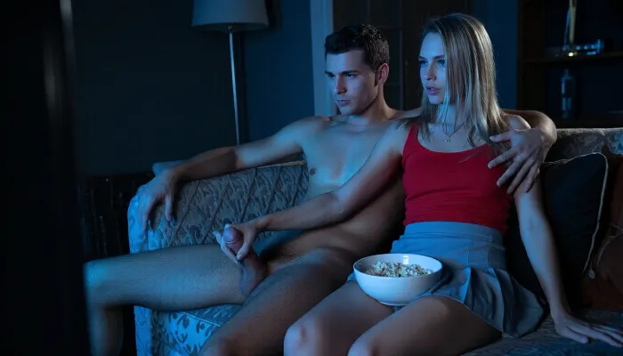 A cinematic, dimly lit indoor scene at night. A young, fit, nude man and a blonde woman sitting upright close together on a plush fabric couch, watching television. The woman is wearing a red tank top and light grey pleated mini skirt, holding a white bowl filled with popcorn on her lap, while her hand reaches down, touching the man's erect penis. The man has one arm around the woman's shoulder, the other arm rests relaxed on the back of the sofa. The man is completely nude, sitting beside her with a focused expression. The room is dark, illuminated only by the cool, flickering blue light from an off-screen television, casting dramatic shadows and highlights on their skin and faces. Photorealistic, high detail, shallow depth of field, movie still aesthetic, dramatic lighting.