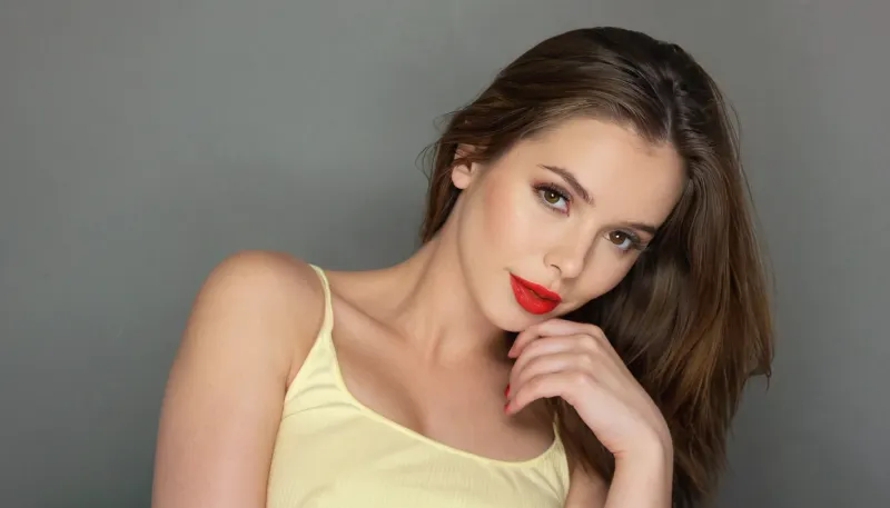 The woman in the image has a light tan complexion. Her brown hair is shoulder-length and appears straight. She is wearing a pale yellow top. Her lips are painted with a vibrant red lipstick, and there is a light touch of blush on her cheeks. Her expression is relaxed, with her hand resting gently on her chin. Her eyes appear to be a light brown color. Her gaze is directed slightly off to the viewer's right.