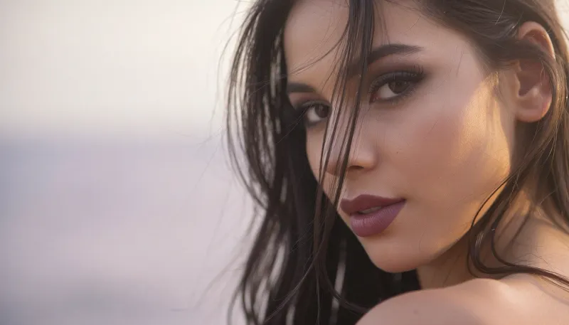 An intimate closeup portrait of a Latina woman with an ethereal, soft-focus aesthetic, defined by diffused light. The Mexican woman's long elegant dark hair falls loosely across her face and obscures her sultry facial features and coyly hiding one eye behind the thick cascade of luxurious hair.
Her thick feathery eyelashes and dark smokey makeup highlight her large sensual eyes.
Her soft full lips are a dark matte berry with visible pillowy creases. She has prominent sharp cheekbones and huge expressive eyes locked on the viewer in a sensual adoring gaze.

The scene is enveloped in a powerful but diffused golden-hour glow from the side, which creates a luminous light bloom on her skin, dissolving sharp edges in an ethereal haze. The background is a very blurred, dreamlike pastoral pastel lavender-gray coastal sky, soft and bright. Soft focus on her luscious kissable lips.
The overall mood is an intimate serene sensuality. Shallow depth of field, subtle film grain, light leaks, vintage analog lens. This is a high-key photograph.
The subject is anchored to the right side of the image, using the rule-of-thirds compositional technique.