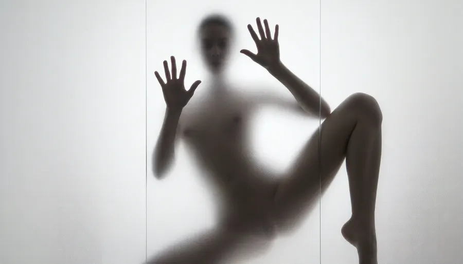 Ultra photorealistic minimalist monochrome fine art photography, vertical composition, mysterious adult female silhouette 28 years old standing behind heavily frosted translucent glass, entire image dominated by luminous white and pale gray tones, figure positioned almost exactly in the center, body visible only as a soft charcoal shadow through condensation and diffused glass texture, no facial detail readable, head slightly inclined, shoulders narrow, torso softly defined, natural feminine proportions, blurred hip and waist transitions, absolutely no sharp body edges, both arms raised forward toward the glass, both flat inner palms pressed firmly against the frosted pane, fingers naturally spread, the two hand contact areas are the only elements in the entire image rendered razor sharp, crisply outlined, perfectly defined and hyperdetailed, individual fingers, fingertips, palm creases, subtle skin texture, fine lines, pressure points and delicate condensation displacement around each hand clearly visible, faint moisture halos surrounding the palms, realistic contact between warm skin and cold glass, while wrists, arms, body, head and legs immediately dissolve back into heavy diffusion and soft blur, thick atmospheric frosting obscuring all other anatomical detail, one leg straight and weight bearing, the other knee bent inward creating an elegant asymmetrical S shaped silhouette, brightest values surrounding the figure and gradually fading toward the center, extremely soft optical blooming, subtle vertical streaks and uneven translucency across the glass, barely perceptible shadows around the feet, dreamy ethereal isolation, no visible room details, no hard light source, broad diffuse backlighting from behind the glass, very low contrast, milky whites, pearl gray midtones and soft graphite silhouette, strong selective focus exclusively on the palms against the pane, everything else intentionally defocused, slight vintage film softness, minimal grain, 85 mm lens compression, shallow tonal depth, haunting anonymous presence, museum quality abstract photographic realism