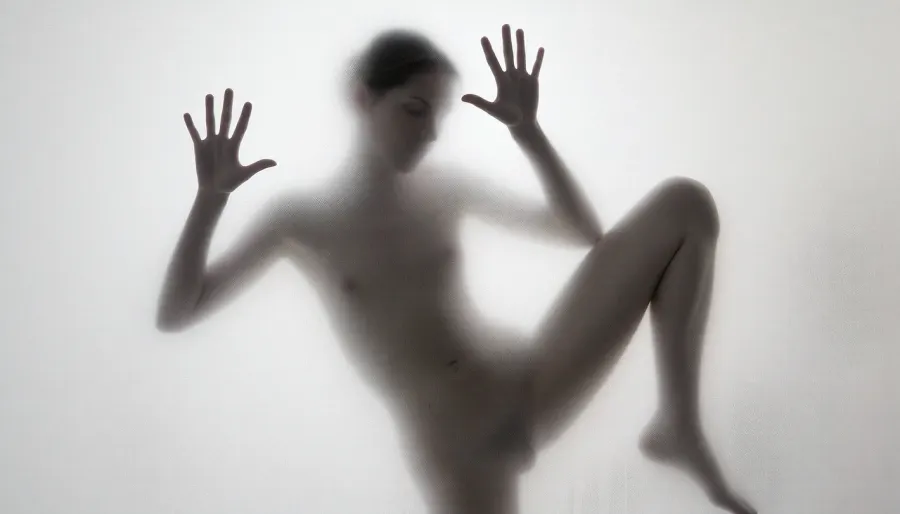 Ultra photorealistic minimalist monochrome fine art photography, vertical composition, mysterious adult female silhouette 28 years old standing behind heavily frosted translucent glass, entire image dominated by luminous white and pale gray tones, figure positioned almost exactly in the center, body visible only as a soft charcoal shadow through condensation and diffused glass texture, no facial detail readable, head slightly inclined, shoulders narrow, torso softly defined, natural feminine proportions, blurred hip and waist transitions, absolutely no sharp body edges, both arms raised forward toward the glass, both flat inner palms pressed firmly against the frosted pane, fingers naturally spread, the two hand contact areas are the only elements in the entire image rendered razor sharp, crisply outlined, perfectly defined and hyperdetailed, individual fingers, fingertips, palm creases, subtle skin texture, fine lines, pressure points and delicate condensation displacement around each hand clearly visible, faint moisture halos surrounding the palms, realistic contact between warm skin and cold glass, while wrists, arms, body, head and legs immediately dissolve back into heavy diffusion and soft blur, thick atmospheric frosting obscuring all other anatomical detail, one leg straight and weight bearing, the other knee bent inward creating an elegant asymmetrical S shaped silhouette, brightest values surrounding the figure and gradually fading toward the center, extremely soft optical blooming, subtle vertical streaks and uneven translucency across the glass, barely perceptible shadows around the feet, dreamy ethereal isolation, no visible room details, no hard light source, broad diffuse backlighting from behind the glass, very low contrast, milky whites, pearl gray midtones and soft graphite silhouette, strong selective focus exclusively on the palms against the pane, everything else intentionally defocused, slight vintage film softness, minimal grain, 85 mm lens compression, shallow tonal depth, haunting anonymous presence, museum quality abstract photographic realism